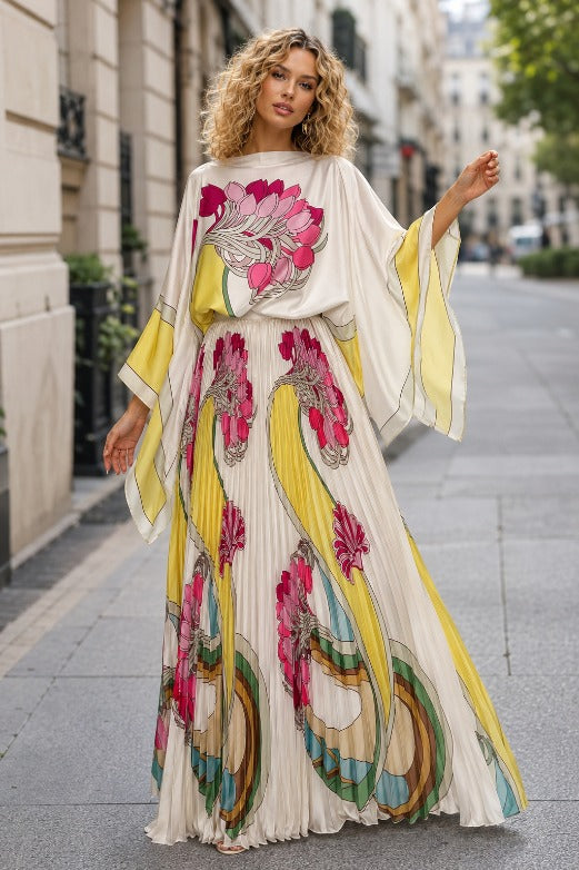 Woman wearing an ivory floral pleated maxi dress with fuchsia and yellow print and flowing cape sleeves